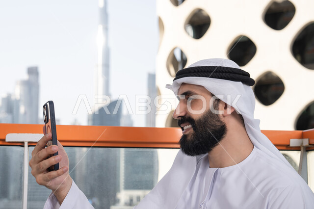 Keeping photos and memorable moments, taking a commemorative selfie with a mobile device with gestures of happiness and joy, a close-up side shot of an Emirati Gulf Arab man wearing a kandura and ghutrah sitting in a café holding a mobile phone in his hand, using a modern and advanced technological device.