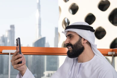 الاحتفاظ بالصور واللحظات التذكارية, التقاط صورة سيلفي تذكارية عبر جهاز الجوال بإيماءات السعادة والسرور،صورة مقربة من الجانب لرجل عربي خليجي اماراتي يرتدي الكندورة و الغتره يجلس فى المقهي يمسك بيده الهاتف المحمول، استخدام جهاز تقني حديث ومتطور