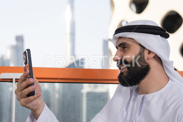 كوفي شوب راقي بالامارات,الاحتفاظ باللحظات السعيدة، التقاط صورة سيلفي تذكارية عبر جهاز الجوال بإيماءات السعادة والسرور، صورة مقربة من الجانب لرجل عربي خليجي اماراتي يرتدي الكندورة و الغتره يجلس فى المقهي يمسك بيده الهاتف المحمول، استخدام جهاز تقني حديث ومتطور