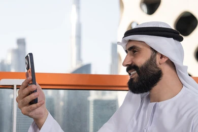 كوفي شوب راقي بالامارات,الاحتفاظ باللحظات السعيدة، التقاط صورة سيلفي تذكارية عبر جهاز الجوال بإيماءات السعادة والسرور، صورة مقربة من الجانب لرجل عربي خليجي اماراتي يرتدي الكندورة و الغتره يجلس فى المقهي يمسك بيده الهاتف المحمول، استخدام جهاز تقني حديث ومتطور