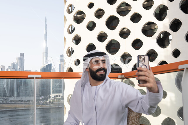 قضاء أوقات ممتعة,الاحتفاظ باللحظات السعيدة، التقاط صورة سيلفي تذكارية عبر جهاز الجوال بإيماءات السعادة والسرور، صورة مقربة لرجل عربي خليجي اماراتي يرتدي الكندورة و الغتره يجلس فى المقهي يمسك بيده الهاتف المحمول، استخدام جهاز تقني حديث ومتطور