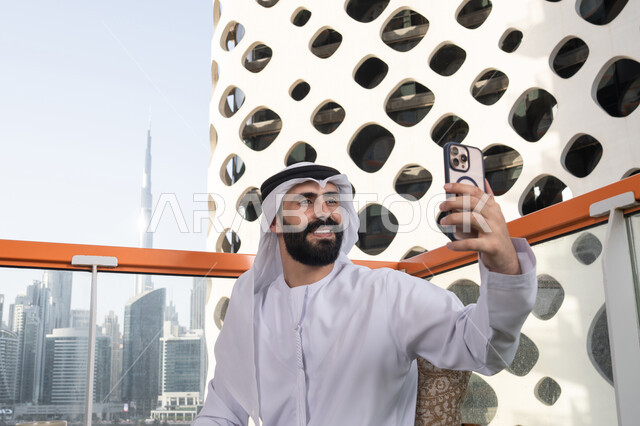 التسلية والترفيه بالعطلات, الاحتفاظ باللحظات السعيدة، التقاط صورة سيلفي تذكارية عبر جهاز الجوال بإيماءات السعادة والسرور، صورة مقربة لرجل عربي خليجي اماراتي يرتدي الكندورة و الغتره يجلس فى المقهي يمسك بيده الهاتف المحمول، استخدام جهاز تقني حديث ومتطور