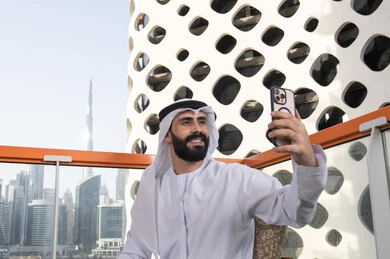 التسلية والترفيه بالعطلات, الاحتفاظ باللحظات السعيدة، التقاط صورة سيلفي تذكارية عبر جهاز الجوال بإيماءات السعادة والسرور، صورة مقربة لرجل عربي خليجي اماراتي يرتدي الكندورة و الغتره يجلس فى المقهي يمسك بيده الهاتف المحمول، استخدام جهاز تقني حديث ومتطور