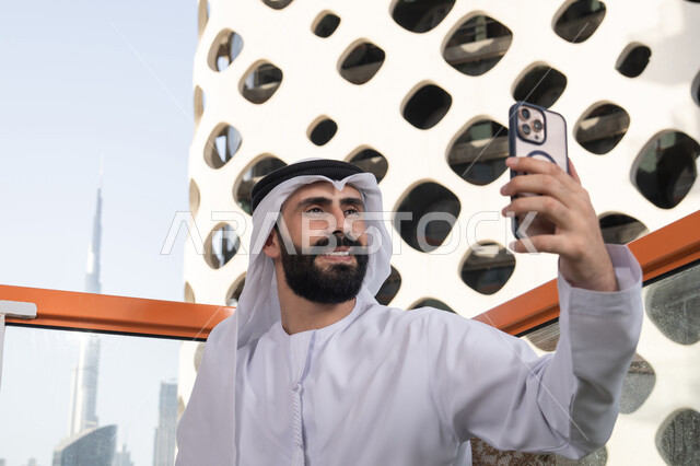 Gestures of happiness and joy, preserving happy moments, taking a commemorative selfie with a mobile device, a close-up image of an Arab Gulf Emirati man wearing a kandura and ghutrah sitting in a café holding a mobile phone in his hand, using a modern and advanced technological device.