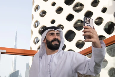 ايماءات السعادة و السرور , الاحتفاظ باللحظات السعيدة، التقاط صورة سيلفي تذكارية عبر جهاز الجوال ، صورة مقربة لرجل عربي خليجي اماراتي يرتدي الكندورة و الغتره يجلس فى المقهي يمسك بيده الهاتف المحمول، استخدام جهاز تقني حديث ومتطور