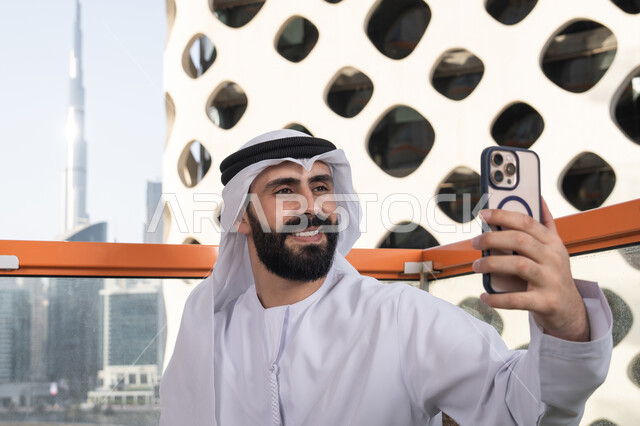 توثيق اللقطات الشخصية التذكارية, الاحتفاظ باللحظات السعيدة، ايماءات السعادة والسرور، صورة مقربة لرجل عربي خليجي اماراتي يرتدي الكندورة و الغتره يجلس فى المقهي يمسك بيده الهاتف المحمول، استخدام جهاز تقني حديث ومتطور