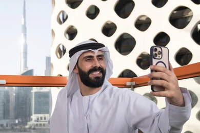 Documenting personal commemorative snapshots, preserving happy moments, gestures of joy and happiness, a close-up image of an Emirati Gulf Arab man wearing a kandura and ghutrah sitting in a café holding a mobile phone, using a modern and advanced technological device.