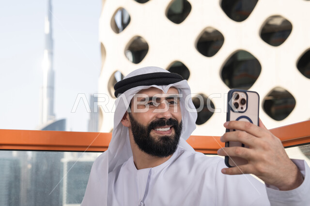 التقاط صورة سيلفي تذكارية عبر جهاز الجوال بإيماءات السعادة والسرور, الاحتفاظ باللحظات السعيدة، صورة مقربة لرجل عربي خليجي اماراتي يرتدي الكندورة و الغتره  يجلس فى المقهي يمسك بيده الهاتف المحمول، استخدام جهاز تقني حديث ومتطور