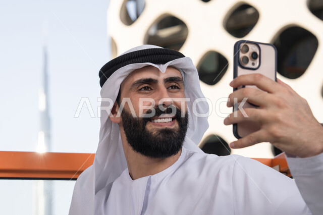 الاحتفاظ باللحظات السعيدة، التقاط صورة سيلفي تذكارية عبر جهاز الجوال بإيماءات السعادة والسرور، صورة مقربة لرجل عربي خليجي اماراتي يرتدي الكندورة و الغتره يجلس فى المقهي يمسك بيده الهاتف المحمول، استخدام جهاز تقني حديث ومتطور