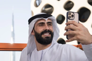 الاحتفاظ باللحظات السعيدة، التقاط صورة سيلفي تذكارية عبر جهاز الجوال بإيماءات السعادة والسرور، صورة مقربة لرجل عربي خليجي اماراتي يرتدي الكندورة و الغتره يجلس فى المقهي يمسك بيده الهاتف المحمول، استخدام جهاز تقني حديث ومتطور