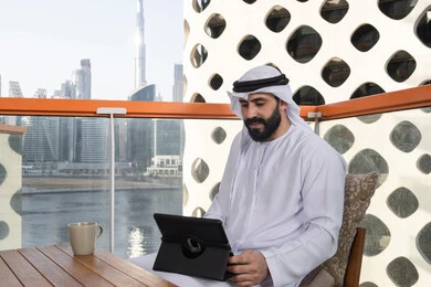القيام بمقابلات وإجتماعات العمل أونلاين,التطور التكنولوجي والتقني في مجالات العمل عن بعد، الدردشة مع الأهل والأصدقاء، رجل عربي خليجي اماراتي يرتدي الكندورة و الغترة البيضاء يجلس فى المقهي و يحمل بيده جهاز التابلت، تصفح مواقع التواصل الاجتماعي، استخدام تطبيقات وبرامج الأجهزة اللوحية