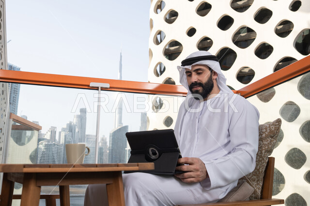 التواصل و الدردشة مع الاقارب,التطور التكنولوجي والتقني في مجالات العمل عن بعد، رجل عربي خليجي اماراتي يرتدي الكندورة و الغترة البيضاء يجلس فى المقهي و يحمل بيده جهاز التابلت، تصفح مواقع التواصل الاجتماعي، استخدام تطبيقات وبرامج الأجهزة اللوحية
