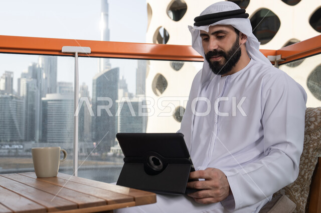 Using a modern technical device to easily browse photos and videos, enjoying a drink, chatting with family and friends, an Emirati Gulf Arab man wearing a kandura and a white ghutrah sitting in a café holding a tablet, browsing social media, using applications and programs for tablets.