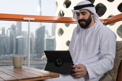 استخدام جهاز تقني حديث لتصفح الصور والفيديوهات بسهولة, الاستمتاع بتناول مشروب, الدردشة مع الأهل والأصدقاء، رجل عربي خليجي اماراتي يرتدي الكندورة و الغترة البيضاء يجلس فى المقهي و يحمل بيده جهاز التابلت، تصفح مواقع التواصل الاجتماعي، استخدام تطبيقات وبرامج الأجهزة اللوحية