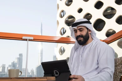 Using the tablet to accomplish work and chat with family and friends, an Arab Gulf Emirati man wearing a kandura and a white ghutrah sits in a café holding a tablet in his hand, browsing social media, using tablet applications and programs, enjoying his favorite drink.