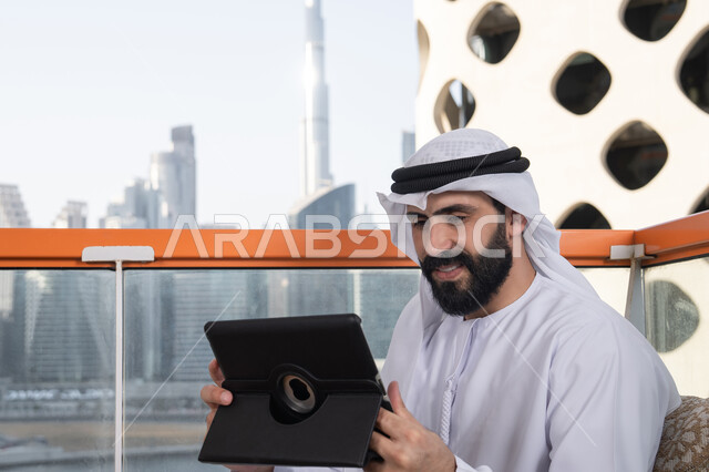 Technological and technical development, chatting with family and friends, an Arab Gulf Emirati man wearing a kandura and a white ghutrah sitting in a café holding a tablet in his hand, browsing social media, using applications and programs for tablets.