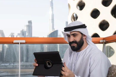 Technological and technical development, chatting with family and friends, an Arab Gulf Emirati man wearing a kandura and a white ghutrah sitting in a café holding a tablet in his hand, browsing social media, using applications and programs for tablets.