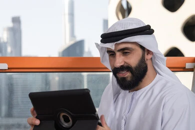 Modern technology in daily life, chatting with family and friends, a close-up image of an Emirati Gulf Arab man wearing a kandura and a white ghutrah sitting in a café holding a tablet device, browsing social media, using applications and programs on tablet devices.