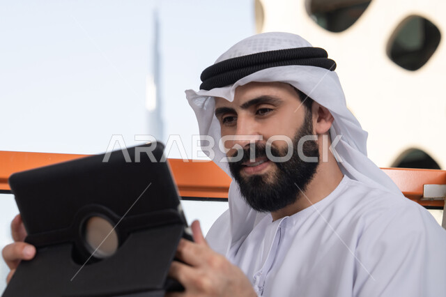 The technological and technical development in the fields of remote work, chatting with family and friends, an Arab Gulf Emirati man wearing a kandura and a white ghutrah sitting in a café holding a tablet in his hand, browsing social media, using applications and programs for tablets.