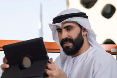 The technological and technical development in the fields of remote work, chatting with family and friends, an Arab Gulf Emirati man wearing a kandura and a white ghutrah sitting in a café holding a tablet in his hand, browsing social media, using applications and programs for tablets.