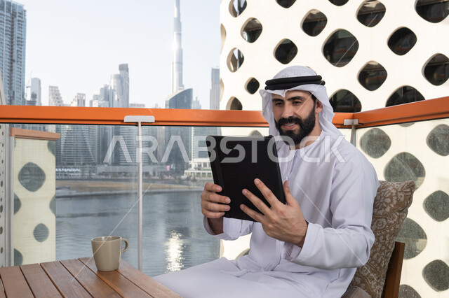 Enjoying drinks, using modern technology and techniques, chatting with family and friends, an Emirati Gulf Arab man wearing a kandura and a white ghutrah holding a tablet in his hand, browsing social media, using applications and programs on tablets.