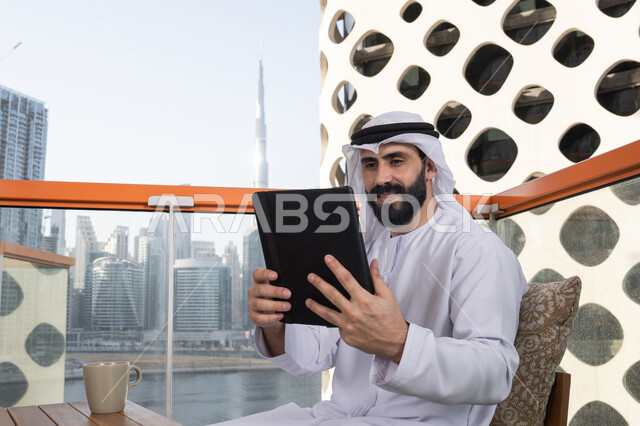 التسويق الالكتروني عن بعد,استعمال التكنولوجيا والتقنيات الحديثة، الدردشة مع الأهل والأصدقاء، رجل عربي خليجي اماراتي يرتدي الكندورة و الغترة البيضاء يجلس فى المقهي و يحمل بيده جهاز التابلت، تصفح مواقع التواصل الاجتماعي، استخدام تطبيقات وبرامج الأجهزة اللوحية