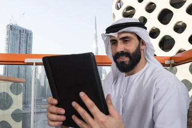 Following the latest news on social media, using technology and modern techniques, chatting with family and friends, an Arab Gulf Emirati man wearing a kandura and a white ghutrah holding a tablet in his hand, browsing social media sites, using applications and programs for tablets.