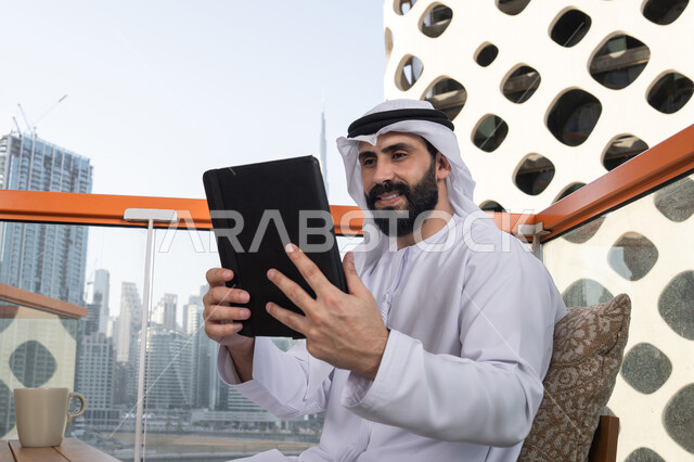 عقد إجتماع عمل عبر الإنترنت, استعمال التكنولوجيا والتقنيات الحديثة، الدردشة مع الأهل والأصدقاء، رجل عربي خليجي اماراتي يرتدي الكندورة و الغترة البيضاء يحمل بيده جهاز التابلت، تصفح مواقع التواصل الاجتماعي، استخدام تطبيقات وبرامج الأجهزة اللوحية