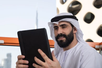 E-marketing, the use of technology and modern techniques, chatting with family and friends, a close-up image of an Arab Gulf Emirati man wearing a kandura and a white ghutrah holding a tablet in his hand, browsing social media sites, using applications and programs on tablets.