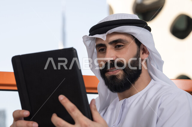 ادارة شؤون الأعمال وتنظيمها عبر الاجهزة التقنية، الدردشة مع الأهل والأصدقاء، صورة مقربة لرجل عربي خليجي اماراتي يرتدي الكندورة و الغترة البيضاء يحمل بيده جهاز التابلت، تصفح مواقع التواصل الاجتماعي، استخدام تطبيقات وبرامج الأجهزة اللوحية
