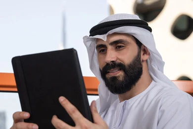 Managing and organizing business affairs through technical devices, chatting with family and friends, a close-up image of an Emirati Gulf Arab man wearing a kandura and a white ghutrah holding a tablet in his hand, browsing social media, using applications and programs on tablets.
