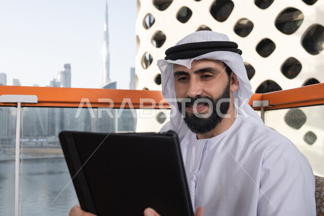 تصفح مواقع التواصل الاجتماعي, استعمال التكنولوجيا والتقنيات الحديثة، الدردشة مع الأهل والأصدقاء، صورة مقربة لرجل عربي خليجي اماراتي يرتدي الكندورة و الغترة البيضاء يحمل بيده جهاز التابلت، استخدام تطبيقات وبرامج الأجهزة اللوحية