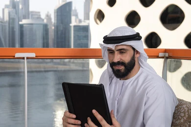 The use of technology and modern techniques, chatting with family and friends, a close-up image of an Arab Gulf Emirati man wearing a kandura and a white ghutrah holding a tablet in his hand, browsing social media, using applications and programs on tablet devices.