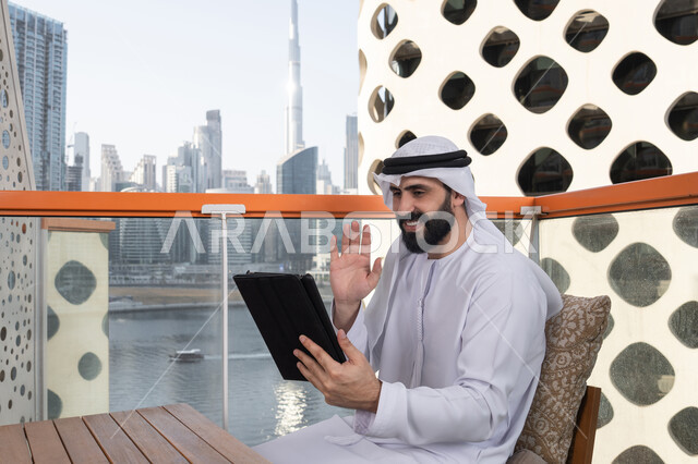 Making a business call, talking to family and friends remotely, browsing the internet and using modern technologies, raising the hand in gestures of greeting and peace, making a video call via a tablet, gestures of joy and happiness, an Emirati Gulf Arab man wearing a kandura and a white ghutrah sitting in a high café overlooking the city.
