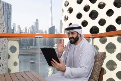 القيام بمكالمة عمل ، التحدث مع الأهل و الأصدقاء عن بعد, تصفح الانترنت واستخدام تقنيات حديثة, رفع الكف للأعلى بايماءات التحية والسلام, إجراء مكالمة فيديو عبر جهاز التابلت, ايماءات السرور والسعادة, رجل عربي خليجي اماراتي يرتدي الكندورة و الغتره البيضاء يجلس في مقهى مرتفع مطل على المدينة