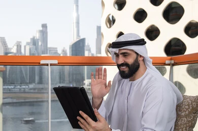 محادثات ودردشات عبر الانترنت ,استخدام تقنيات حديثة, رفع الكف للأعلى بايماءات التحية والسلام, إجراء مكالمة فيديو عبر جهاز التابلت, ايماءات السرور والسعادة, العمل عن بعد من خلال الانترنت, رجل عربي خليجي اماراتي يرتدي الكندورة و الغتره البيضاء يجلس في مقهى مرتفع مطل على المدينة