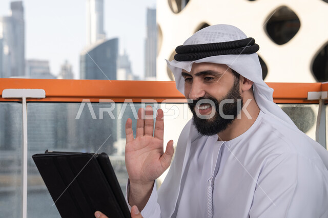 تطور التكنولوجيا وإستخدام الأجهزة التقنية الحديثة, رفع الكف للأعلى بايماءات التحية والسلام, إجراء مكالمة فيديو عبر جهاز التابلت, ايماءات السرور والسعادة, العمل عن بعد من خلال الانترنت, رجل عربي خليجي اماراتي يرتدي الكندورة و الغتره البيضاء يجلس في مقهى مرتفع مطل على المدينة