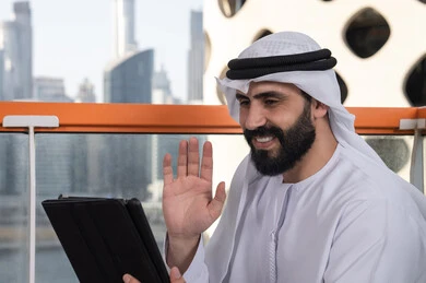 The development of technology and the use of modern technical devices, raising the hand upwards in gestures of greeting and peace, making a video call via a tablet, gestures of joy and happiness, working remotely through the internet, an Arab Gulf Emirati man wearing a kandura and a white ghutrah sitting in a high café overlooking the city.