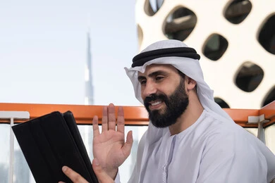 استخدام جهاز تقني حديث ومتطور, رفع الكف للأعلى بايماءات التحية والسلام, إجراء مكالمة فيديو عبر جهاز التابلت, ايماءات السرور والسعادة, العمل عن بعد من خلال الانترنت, رجل عربي خليجي اماراتي يرتدي الكندورة و الغتره البيضاء يجلس في مقهى مرتفع مطل على المدينة