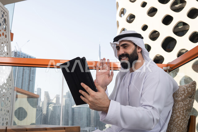 استعمال الجهاز اللوحي في إنجاز العمل, رفع الكف للأعلى بايماءات التحية والسلام, إجراء مكالمة فيديو عبر جهاز التابلت, ايماءات السرور والسعادة, العمل عن بعد من خلال الانترنت, رجل عربي خليجي اماراتي يرتدي الكندورة و الغتره البيضاء يجلس في مقهى مرتفع مطل على المدينة