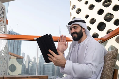 استعمال الجهاز اللوحي في إنجاز العمل, رفع الكف للأعلى بايماءات التحية والسلام, إجراء مكالمة فيديو عبر جهاز التابلت, ايماءات السرور والسعادة, العمل عن بعد من خلال الانترنت, رجل عربي خليجي اماراتي يرتدي الكندورة و الغتره البيضاء يجلس في مقهى مرتفع مطل على المدينة