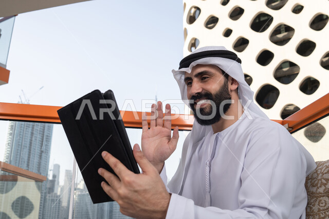 Monitoring and supervising work via a tablet, raising the hand upwards in gestures of greeting and peace, making a video call, gestures of joy and happiness, working remotely through the internet, an Arab Gulf Emirati man wearing a kandura and a white ghutrah sitting in a high café overlooking the city.