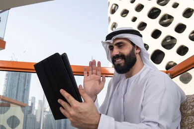 المتابعة والإشراف على العمل عبر جهاز التابلت, رفع الكف للأعلى بايماءات التحية والسلام, إجراء مكالمة فيديو, ايماءات السرور والسعادة, العمل عن بعد من خلال الانترنت, رجل عربي خليجي اماراتي يرتدي الكندورة و الغتره البيضاء يجلس في مقهى مرتفع مطل على المدينة