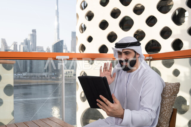 استعمال التكنولوجيا في العمل عن بعد. تصفح الانترنت واستخدام تقنيات حديثة, رفع الكف للأعلى بايماءات التحية والسلام, إجراء مكالمة فيديو عبر جهاز التابلت, ايماءات السرور والسعادة, رجل عربي خليجي اماراتي يرتدي الكندورة و الغتره البيضاء يجلس في مقهى مرتفع مطل على المدينة