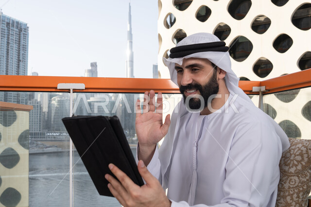 الدردشة مع الأهل والأصحاب, تصفح الانترنت واستخدام تقنيات حديثة, رفع الكف للأعلى بايماءات التحية والسلام, إجراء مكالمة فيديو عبر جهاز التابلت, ايماءات السرور والسعادة, العمل عن بعد من خلال الانترنت, رجل عربي خليجي اماراتي يرتدي الكندورة و الغتره البيضاء يجلس في مقهى مرتفع مطل على المدينة