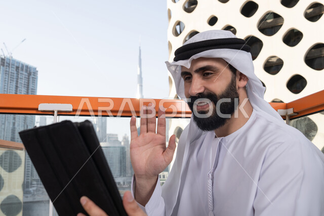 رجل عربي خليجي اماراتي يرتدي الكندورة و الغتره البيضاء يجلس في مقهى مرتفع مطل على المدينة, تصفح الانترنت واستخدام تقنيات حديثة, رفع الكف للأعلى بايماءات التحية والسلام, إجراء مكالمة فيديو عبر جهاز التابلت, ايماءات السرور والسعادة, العمل عن بعد من خلال الانترنت