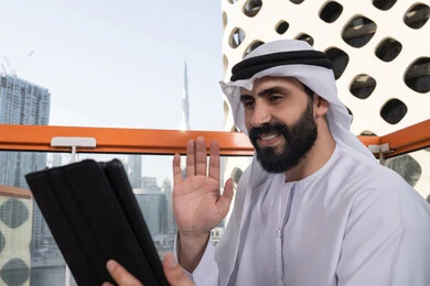 رجل عربي خليجي اماراتي يرتدي الكندورة و الغتره البيضاء يجلس في مقهى مرتفع مطل على المدينة, تصفح الانترنت واستخدام تقنيات حديثة, رفع الكف للأعلى بايماءات التحية والسلام, إجراء مكالمة فيديو عبر جهاز التابلت, ايماءات السرور والسعادة, العمل عن بعد من خلال الانترنت