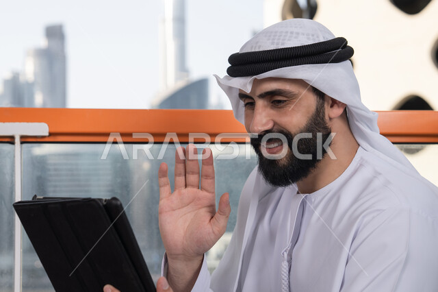 Remote work through the internet, browsing the internet and using modern technologies, raising the hand upwards in gestures of greeting and peace, making a video call via a tablet, gestures of joy and happiness, an Emirati Gulf Arab man wearing a kandura and a white ghutrah sitting in a high café overlooking the city.