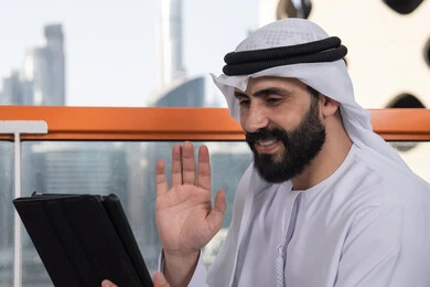 العمل عن بعد من خلال الانترنت, تصفح الانترنت واستخدام تقنيات حديثة, رفع الكف للأعلى بايماءات التحية والسلام, إجراء مكالمة فيديو عبر جهاز التابلت, ايماءات السرور والسعادة, رجل عربي خليجي اماراتي يرتدي الكندورة و الغتره البيضاء يجلس في مقهى مرتفع مطل على المدينة
