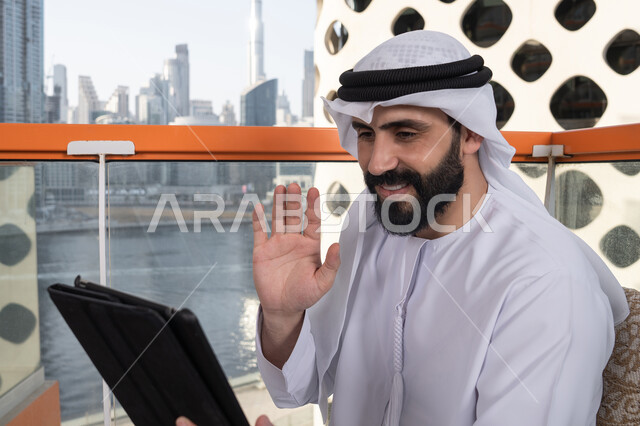 ايماءات السرور والسعادة, تصفح الانترنت واستخدام تقنيات حديثة, رفع الكف للأعلى بايماءات التحية والسلام, إجراء مكالمة فيديو عبر جهاز التابلت, العمل عن بعد من خلال الانترنت, رجل عربي خليجي اماراتي يرتدي الكندورة و الغتره البيضاء يجلس في مقهى مرتفع مطل على المدينة